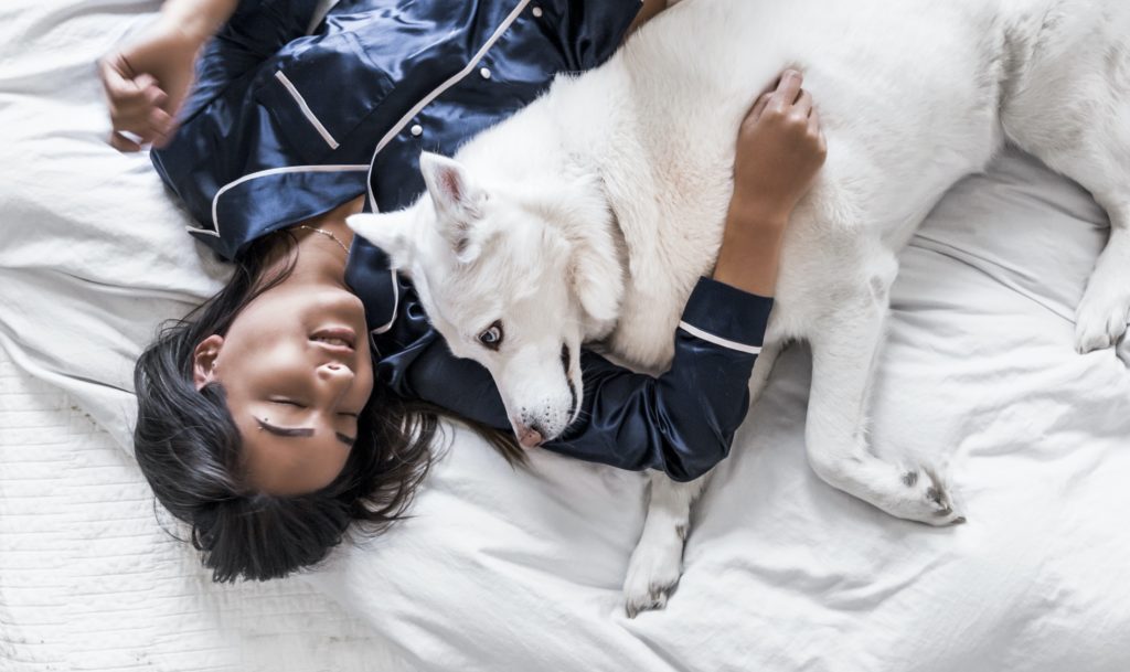 Woman wearing pajamas laying in bed with her arm around a white dog