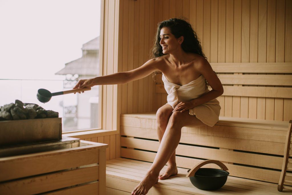 Attractive young woman pouring water onto hot stone in the sauna